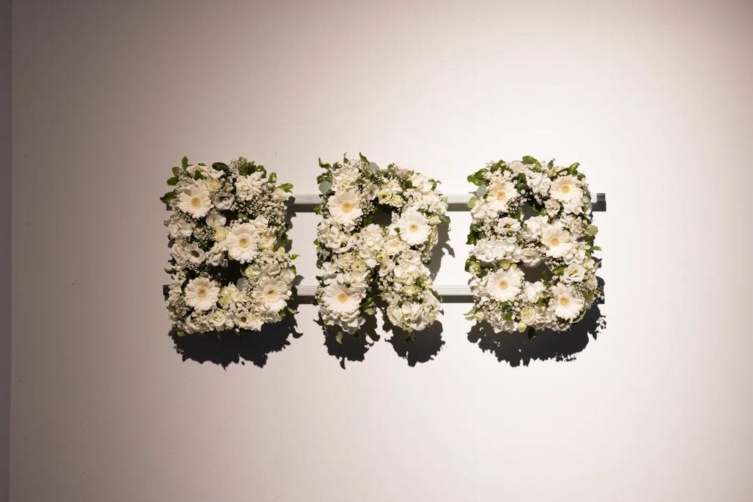 Floral arrangement of white flowers in the letters 'BRB' hang on a clear white studio wall.