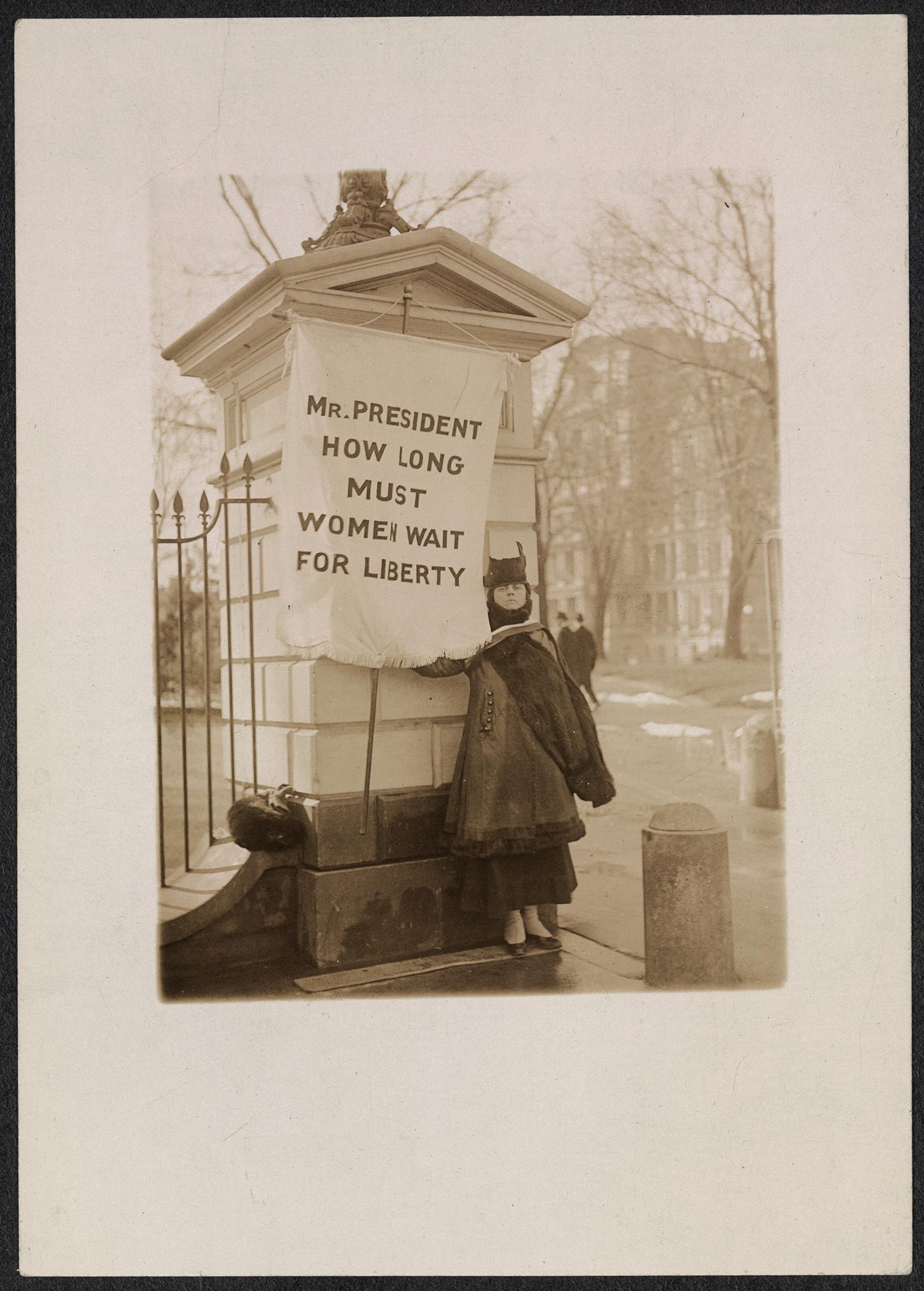 An image of a woman dressed in 1917-style apparel holding a banner reading, "Mr. President, how long must women wait for liberty."