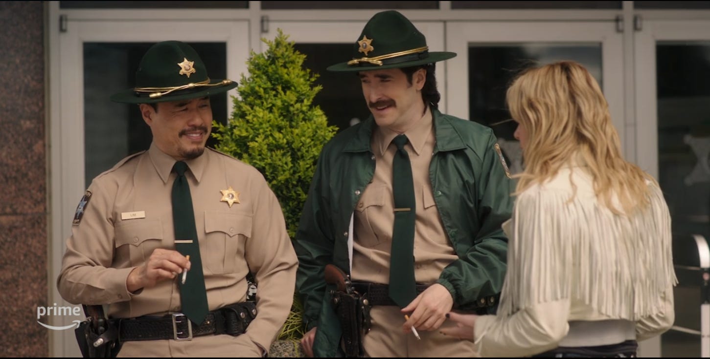 Two American police with 80s facial hair laugh at a young blonde woman in a leather tassel jacket.