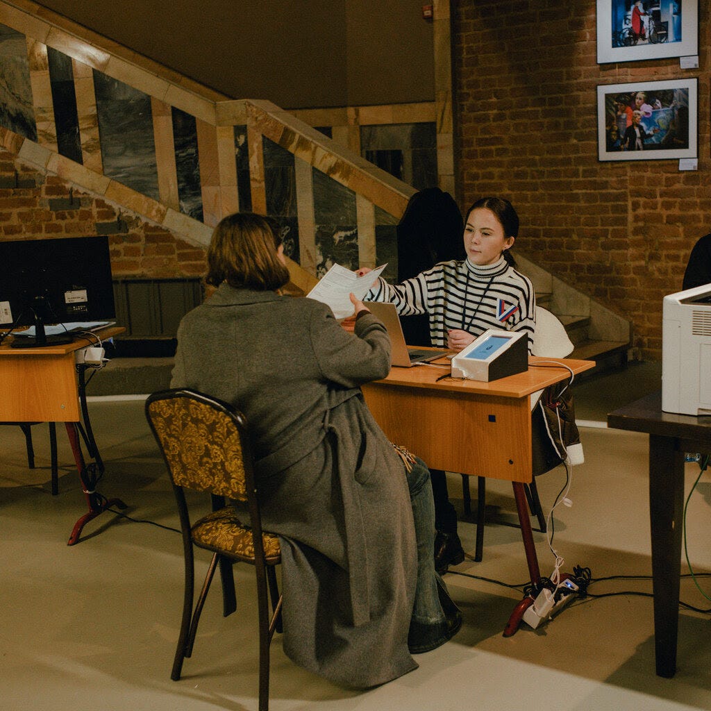 Two women siting on either side of a table inside a brick-walled building pass a paper between them.
