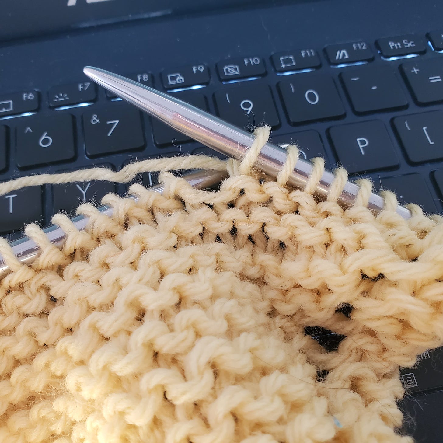 Pale yellow yarn knit into a big garter stitch swatch with metallic circular knitting needles, on a black laptop keyboard