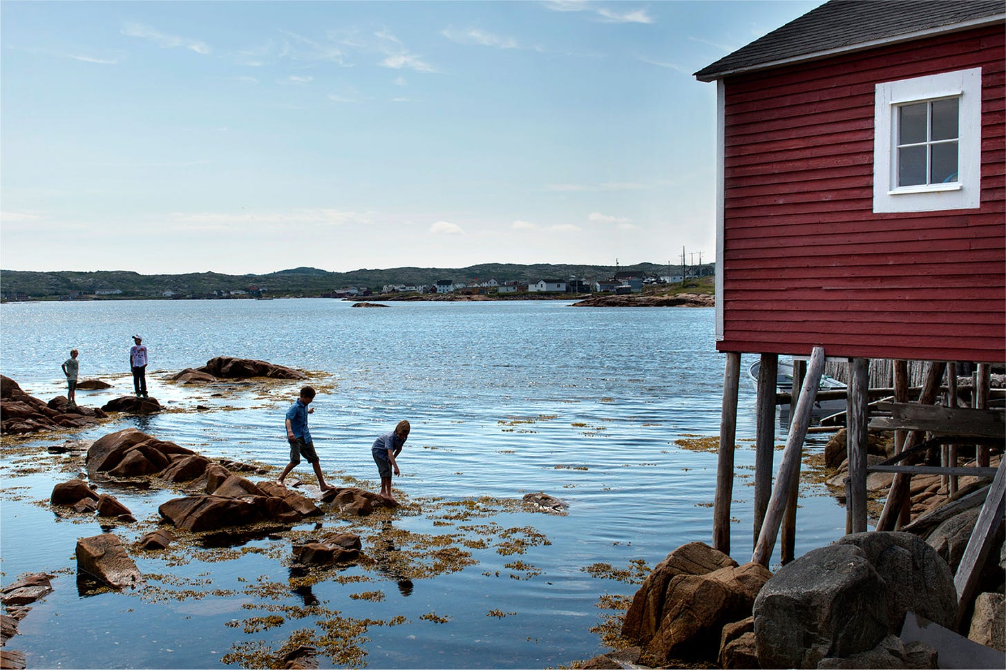 Photograph of four people standing on rocks at the edge of a body of calm blue water; a rustic red building on stilts edges into the frame from the right and other buildings and boats are barely visible in the distance
