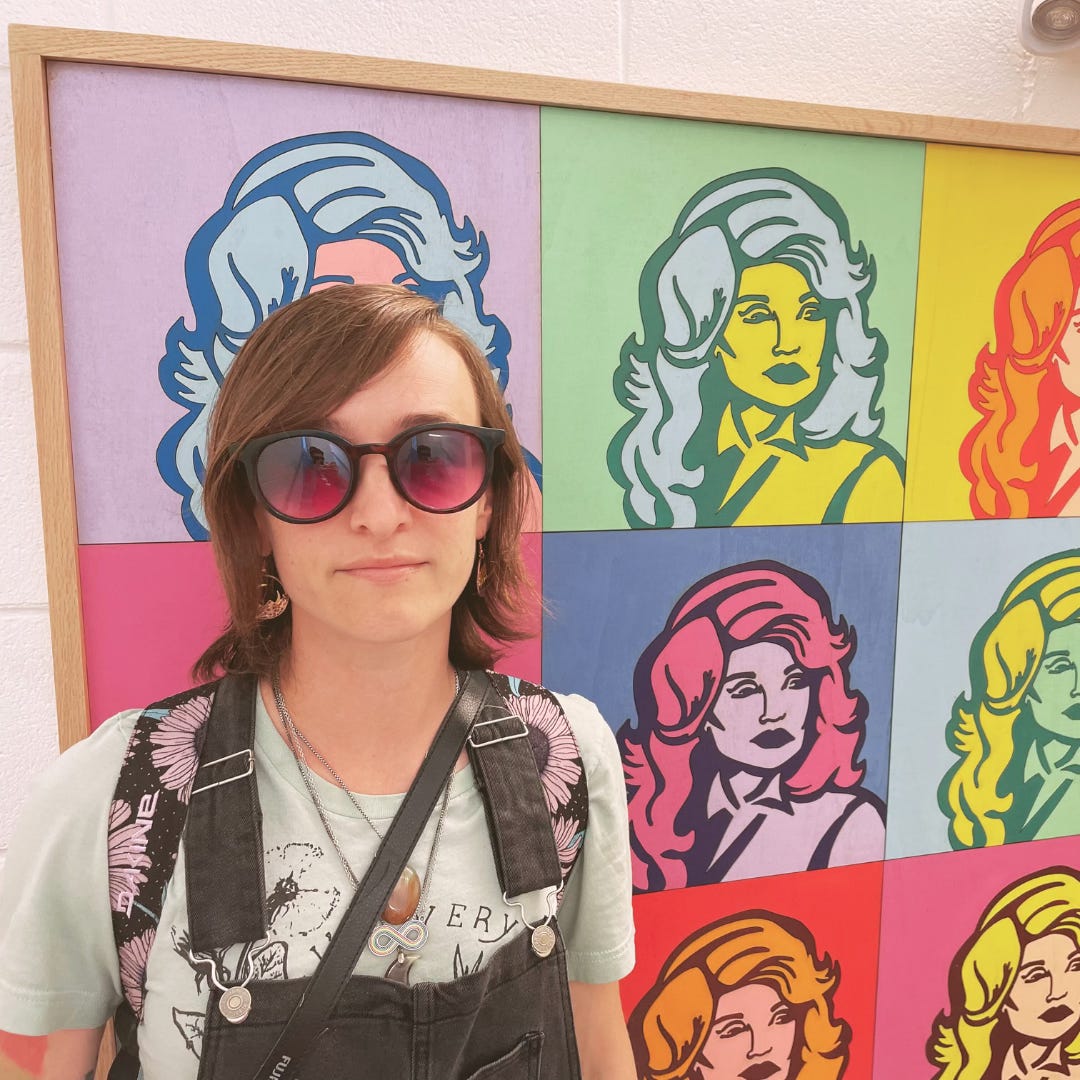 A photograph of Sarah, who wears sunglasses and does not smiling, standing in front of a Pop Art image of Dolly Parton.