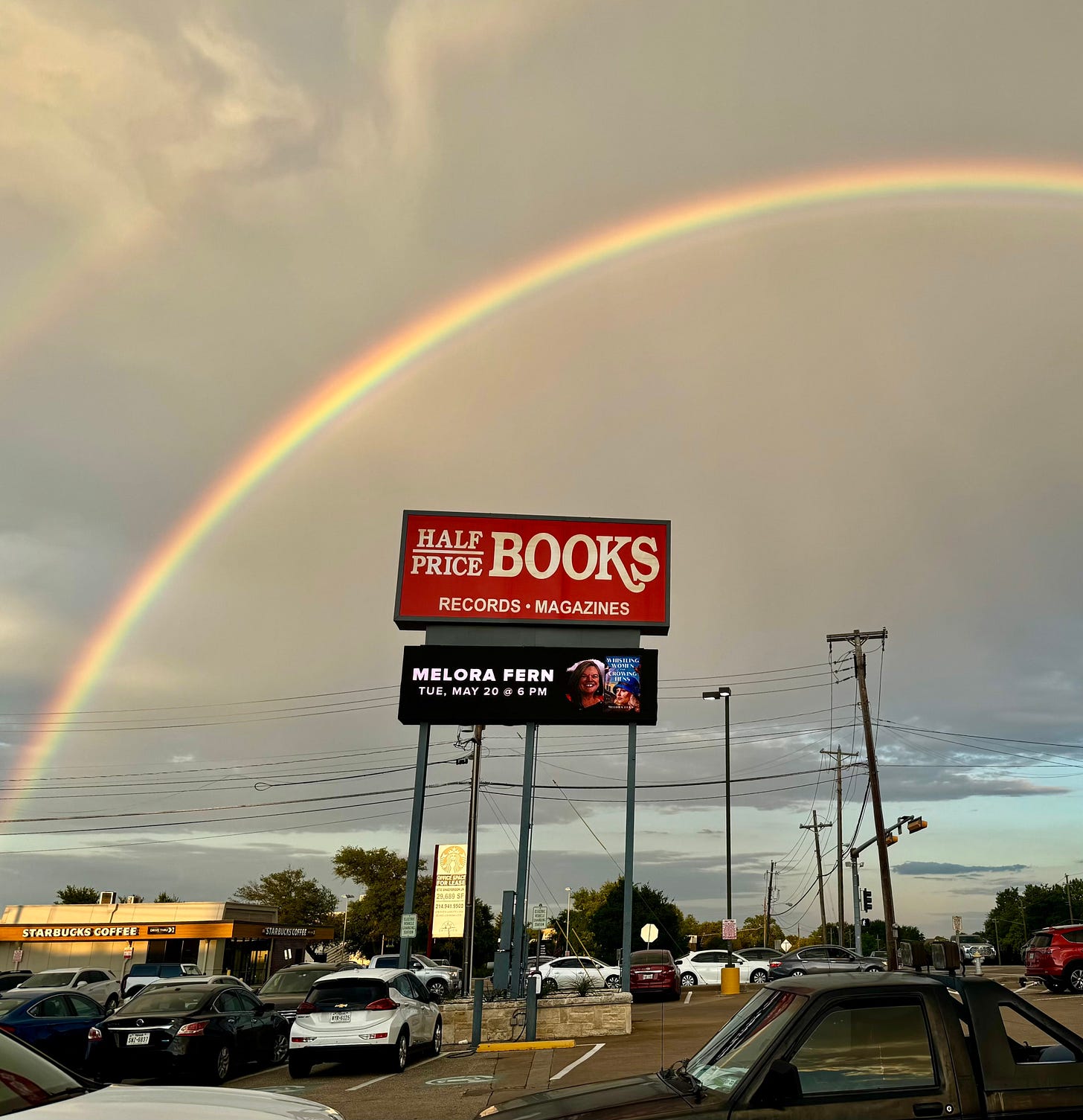 a photo of the half price books sign with Melora Fern and Whistling Women and Crowing Hens featured on the marquee. There is a rainbow in the background