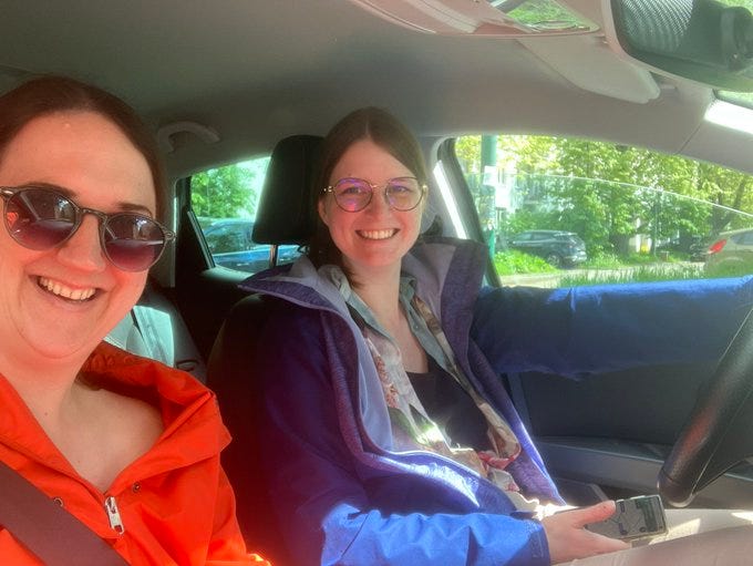 Me wearing my orange jacket and sunnies next to my friend with her glasses on sitting in the car ready to drive.