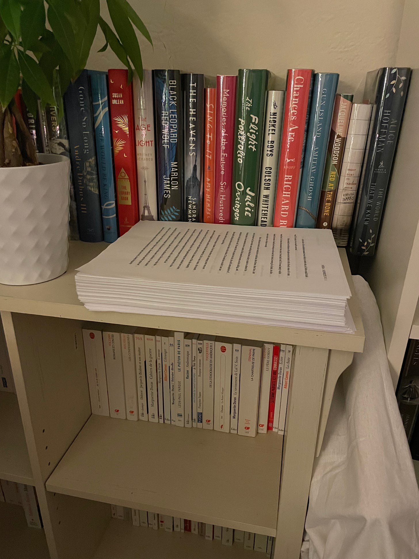 A giant stack of printed paper sits on a white bookshelf next to a number of hardcover books