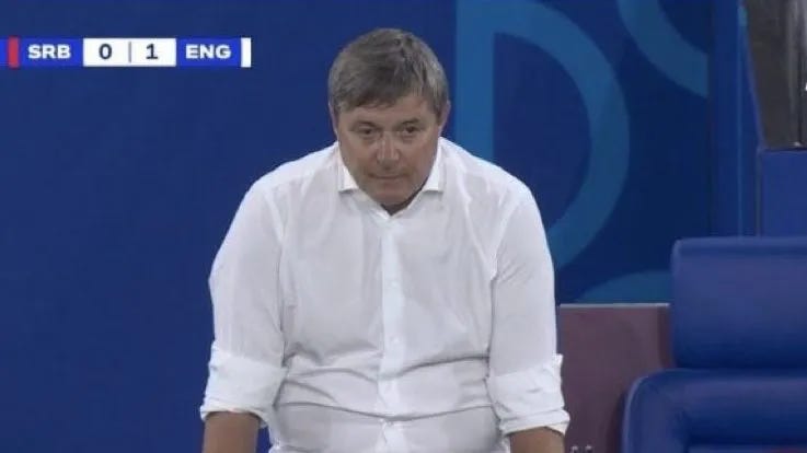 Serbian football coach Dragan Stojkovic Piski, a white man with graying hair and a white button-down shirt with rolled-up sleeves, sitting with slumped shoulders by the side of the soccer field.