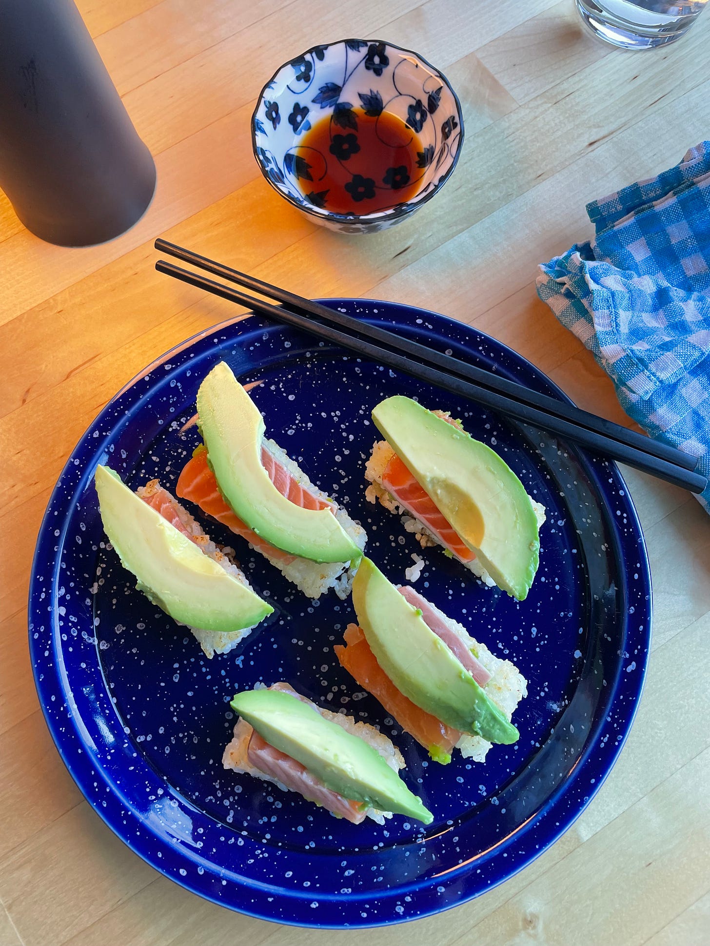 Speckled plate with crispy rice rectangles topped with sliced salmon sashimi and avocado. Some soy sauce and chopsticks nearby.