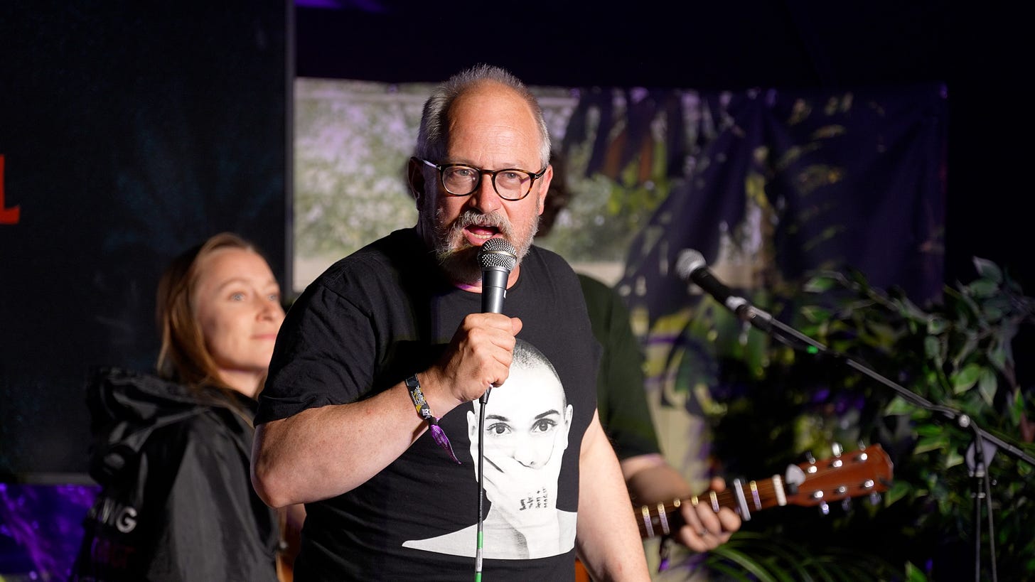 Man on stage holding a microphone wearing a t-shirt with Sinead O'Connor on it