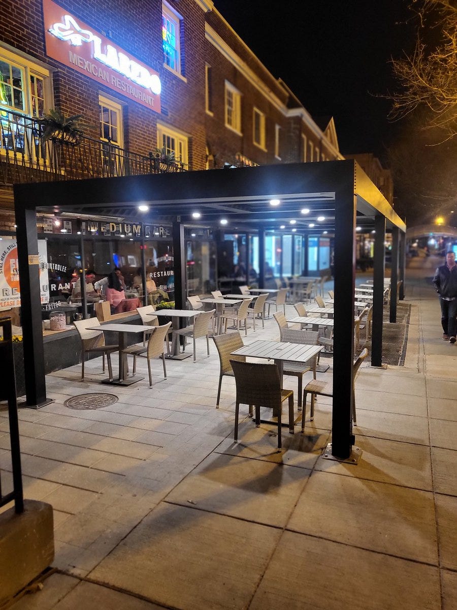 Picture of Medium Rare pergola at night with lights on. The pergola is black with 16 posts and has outdoor seats and tables underneath. It fits with these sidewalk and looks like the perfect spot for a date. In the background you can clearly see the buildings and the Laredo sign. 