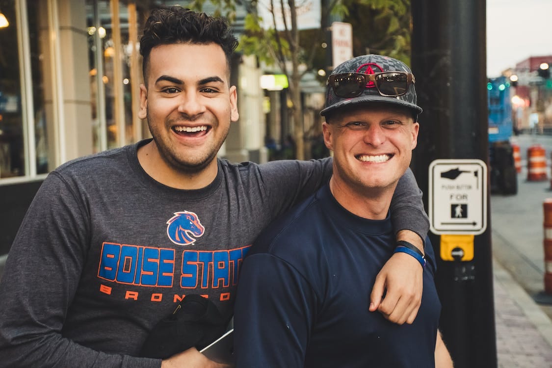 two male friends embracing on an urban street and smiling big for the camera. one is ambigiously poc in a boise state broncos shirt, the other white in a plain blue shirt
