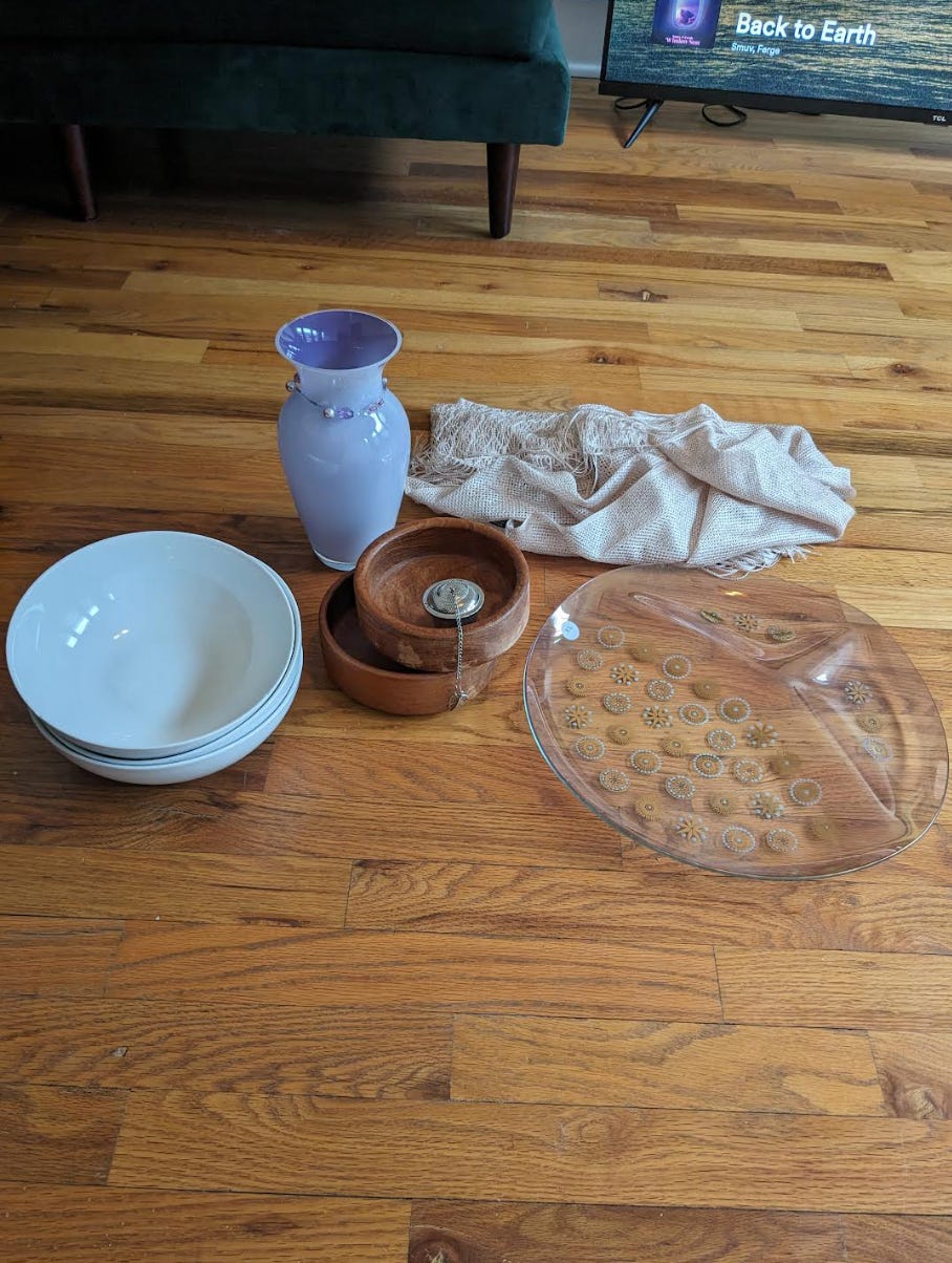 3 white bowls are stacked atop one another, 2 small wooden bowls, one large flat purple/clear plate with gold specks of flower like designs on it, a lavender vase with different shades of purple pearl necklace around the neck of the vase, and a shimmery gold scarf are on the floor of Nathalie's apartment