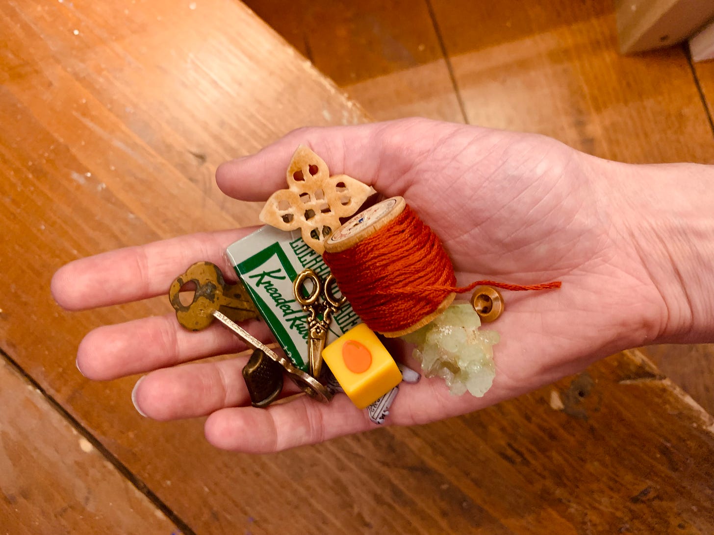 A photo of Gracie's hand over a warm wooden background, holding a small pile of random objects--metal keys, a thimble, a kneaded eraser in it original packaging, a yellow die, some orange embroidery floss, etc. 