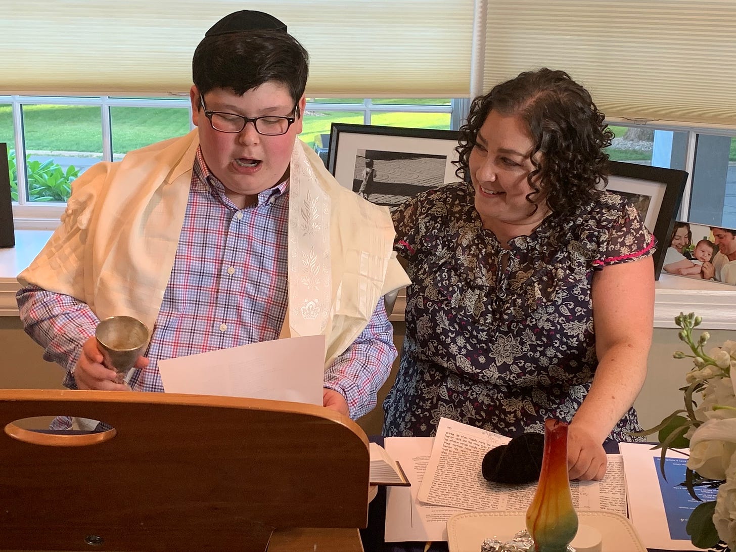 Rafi is a teen boy wearing a kippah and tallit. he sits by a teacher practicing the kiddush, with a kiddush cup in hand