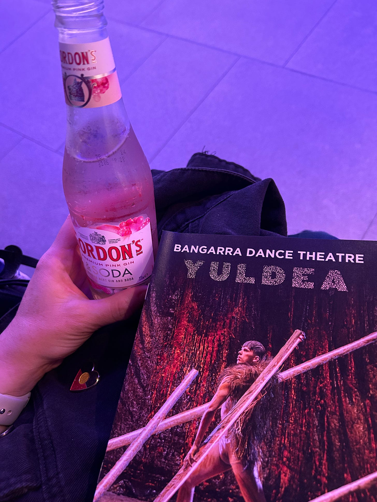 My hand holding a bottle of Gordon's pink gin and soda with the Yuldea program next to it. You can see my black denim jacket underneath and a love heart pin with the colours of the Aboriginal flag.