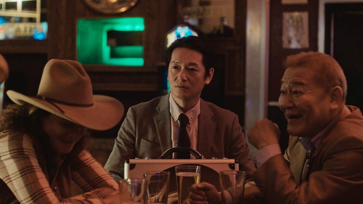 A woman in a cowboy hat is laughing at a joke the older Japanese man across the table is tellling.  A younger Japanese man looks on with a confused expression.