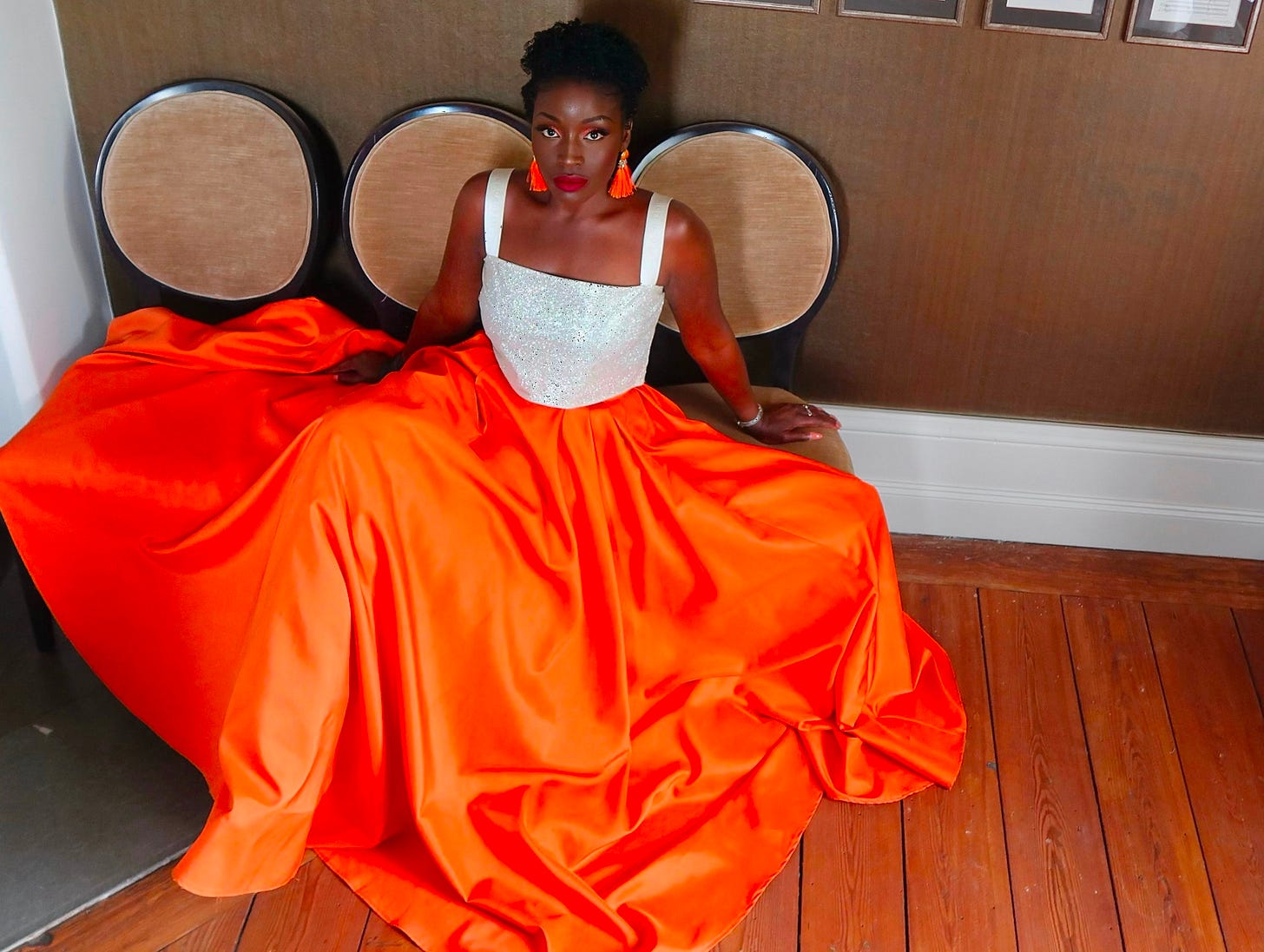 Lovette Jallow in a white and orange gown, seated against a neutral wall with framed text—symbolizing stillness before reflection. Lovette Jallow in a white and orange gown, seated against a neutral wall with framed text—symbolizing stillness before reflection.