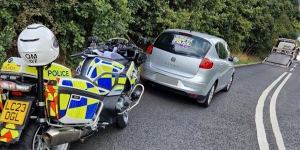 Police motorcycle and seized car parked at the side of a rural road with a transporter lorry in the distance