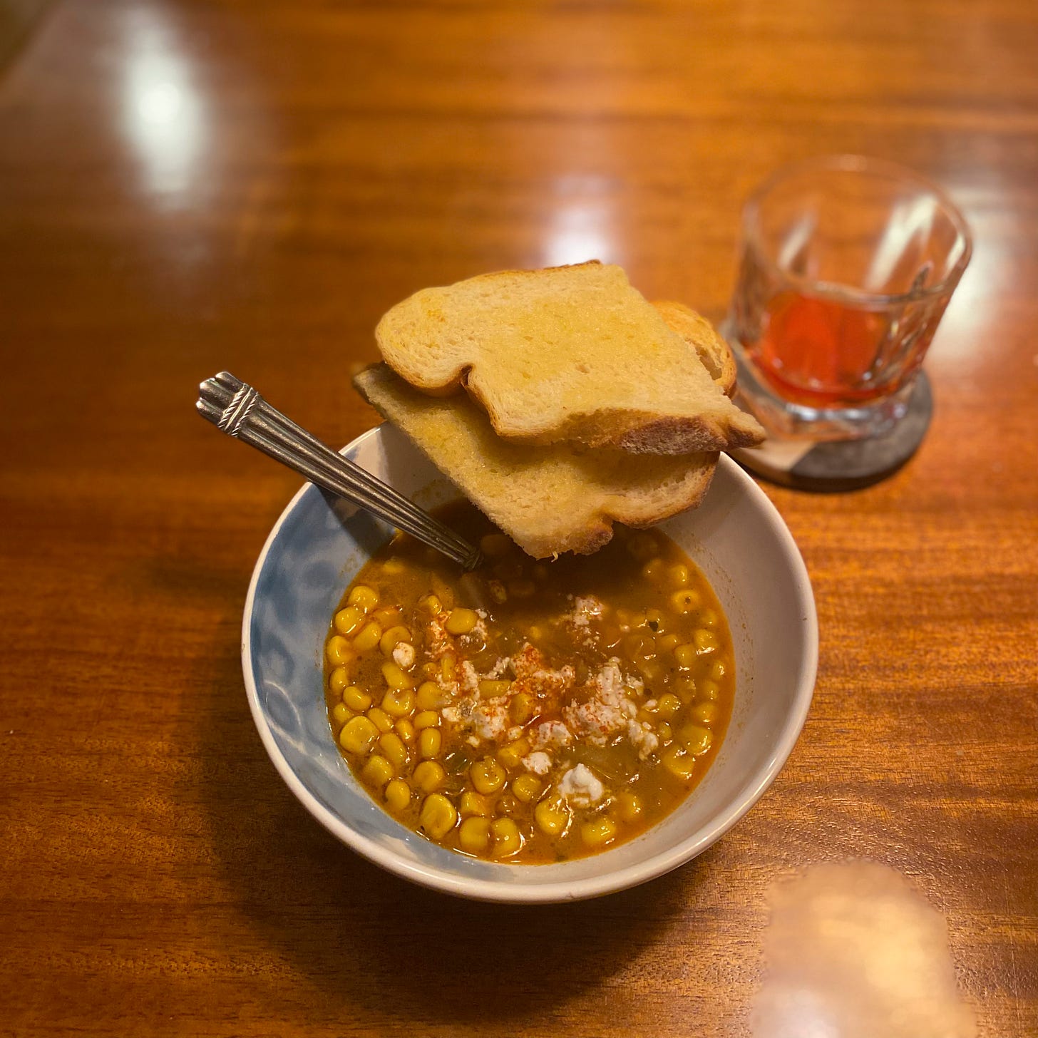 A chunky corn chowder with orangeish broth and crumbles of feta on top. On the edge of the bowl are two halves of a slice of toast, and on a coaster is a mostly-empty rocks glass that had a boulevardier in it.