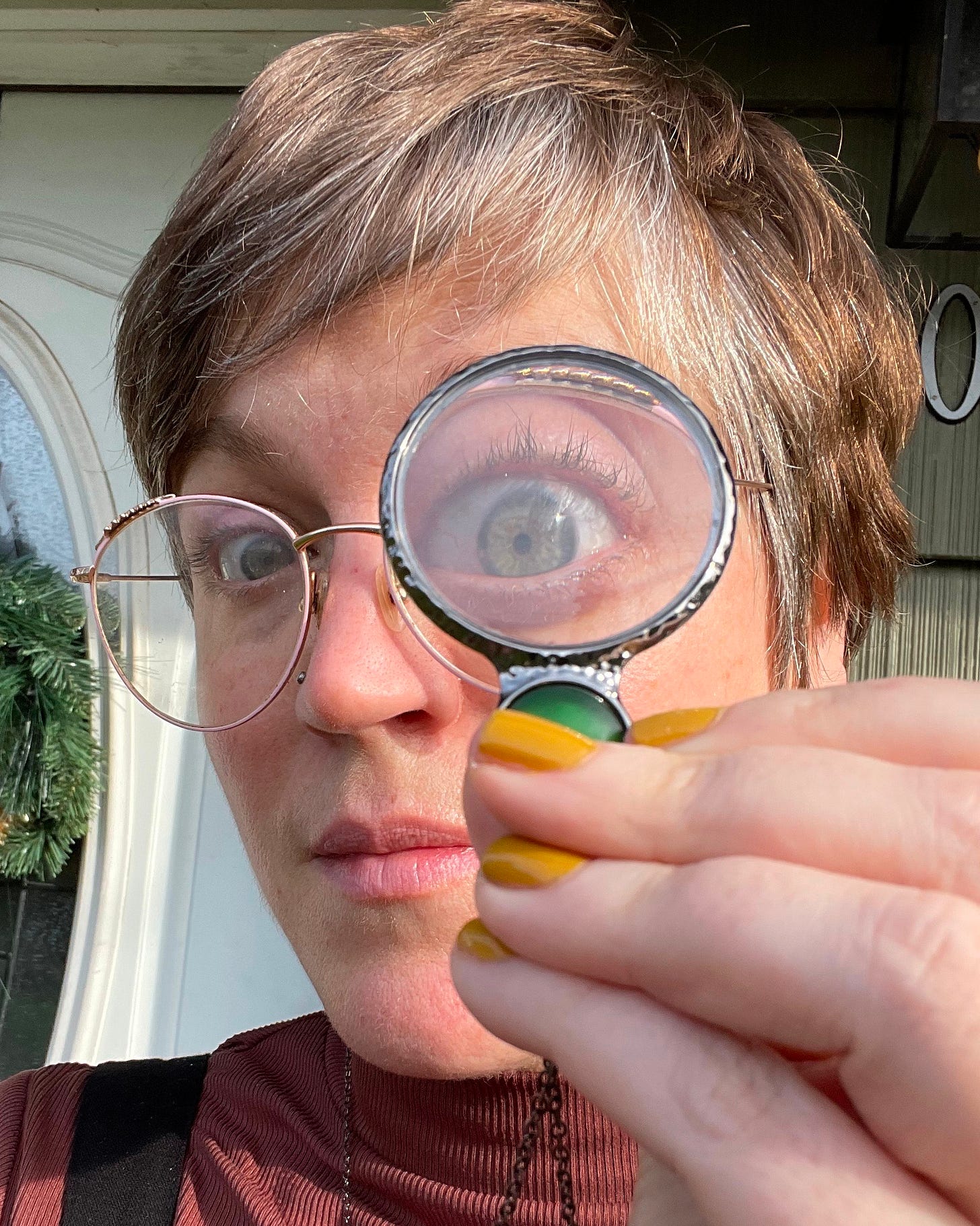 a white person with short brown/gray hair wearing glasses is holding up a magnifying glass to their eye. the glass is attached toa  chain they are wearing around their neck