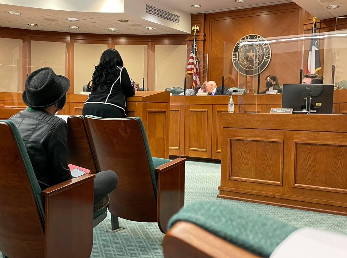 Lyric, a pale skinned nonbinary human in black hat sitting in the stands listening to a bad-ass Black, mother of an Autistic child, with long dark curly hair bravely testifying about the harms of ABA in one of the 2021 Texas legislative meetings. 