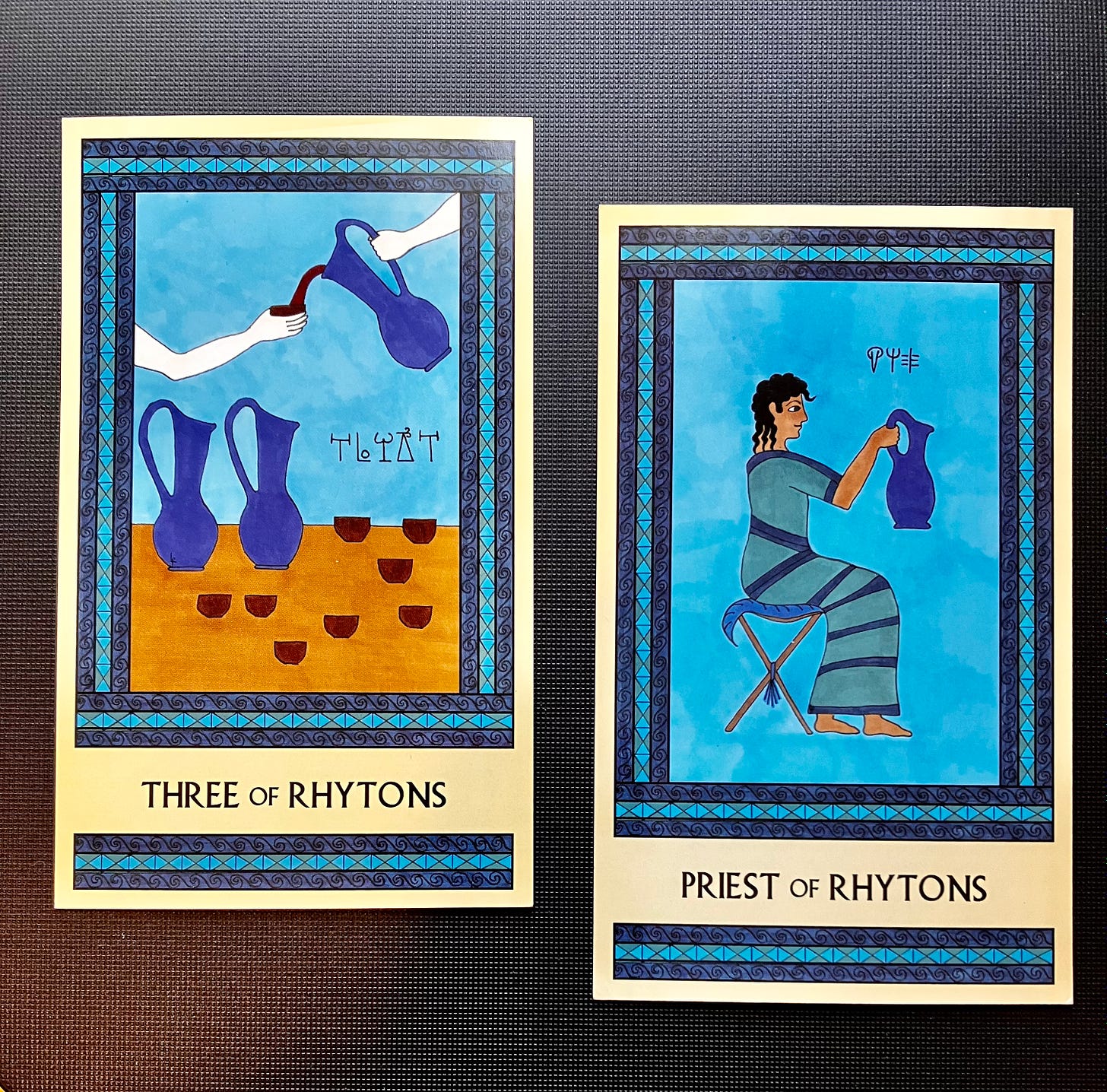 Two Minoan Tarot cards side by side on a black surface. Both cards are in shades of blue. The Three of Rhytons shows a hand holding a pitcher that is pouring wine into another's cup. There are more pitchers and cups on the table. The Priest of Rhytons shows a Minoan man in long robes, seated, facing right and holding up a large blue pitcher.