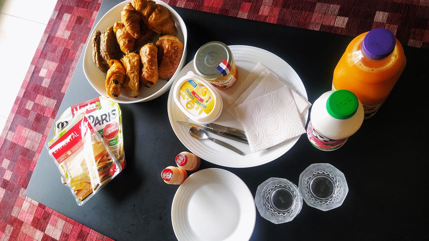 Looking from the top of a black coffee table. There’s a bowl filled with croissants and pain au chocolat, and packets of cheese and ham beside it. Next to it there’s butter, jam, cutlery and napkins on a white dish. To the side there’s two bottles, one of orange juice and another of yogurt. Two probiotic bottles, a smaller dish and two glasses complete the ensemble.