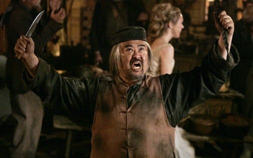 This image shows Mr. Wu (played by Keone Young) holding a knife in his right hand and his Chinese queue (hair braid) in his left hand as he yells up at the off-camera Al Swearengen.