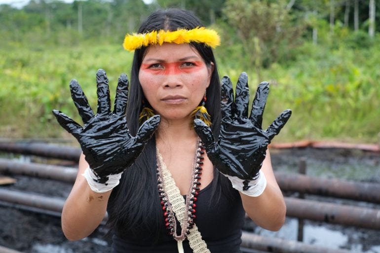 An INemonte Nenquimo, her head crowned in yellow feathers, raises two hands covered in black tar