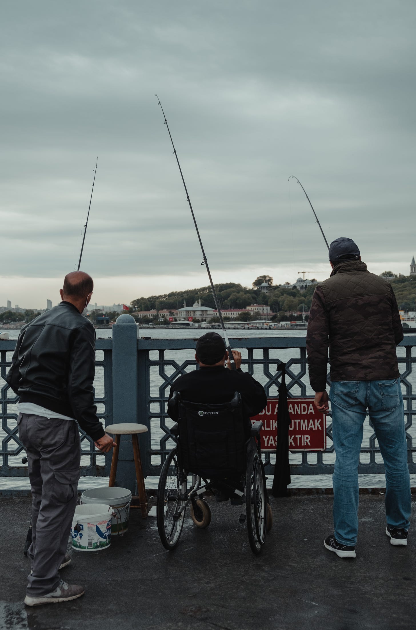 Three men fishing, the middle one is seated in a wheelchair. All of the men have their backs to the camera .