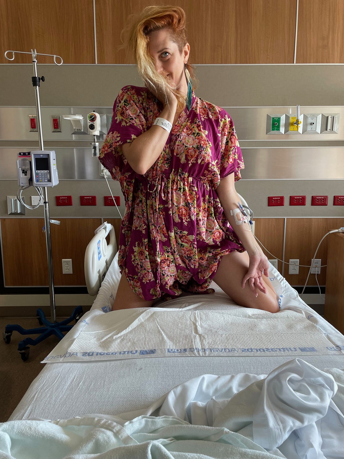 A thin white woman with red hair sits up on her knees on a hospital bed. She peeks out from behind her hair suggestively.