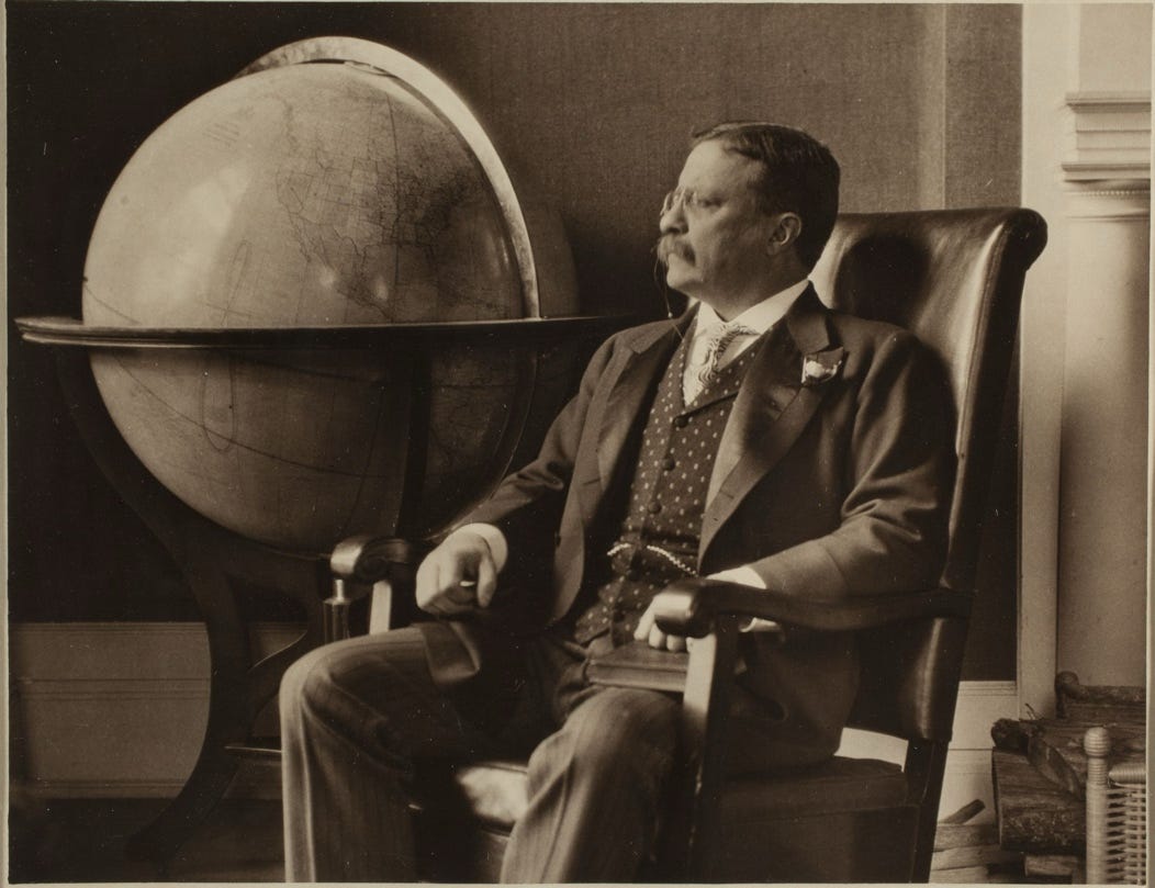 Theodore Roosevelt sits in a chair with a book on his left knee