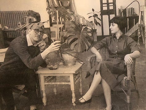 A couple sits on opposite sides of a table with a large plant. The woman has crossed legs and is turned, back straight, looking at the man who pretends to hide behind the plant, leaning over the plant's table. They are in a large studio.