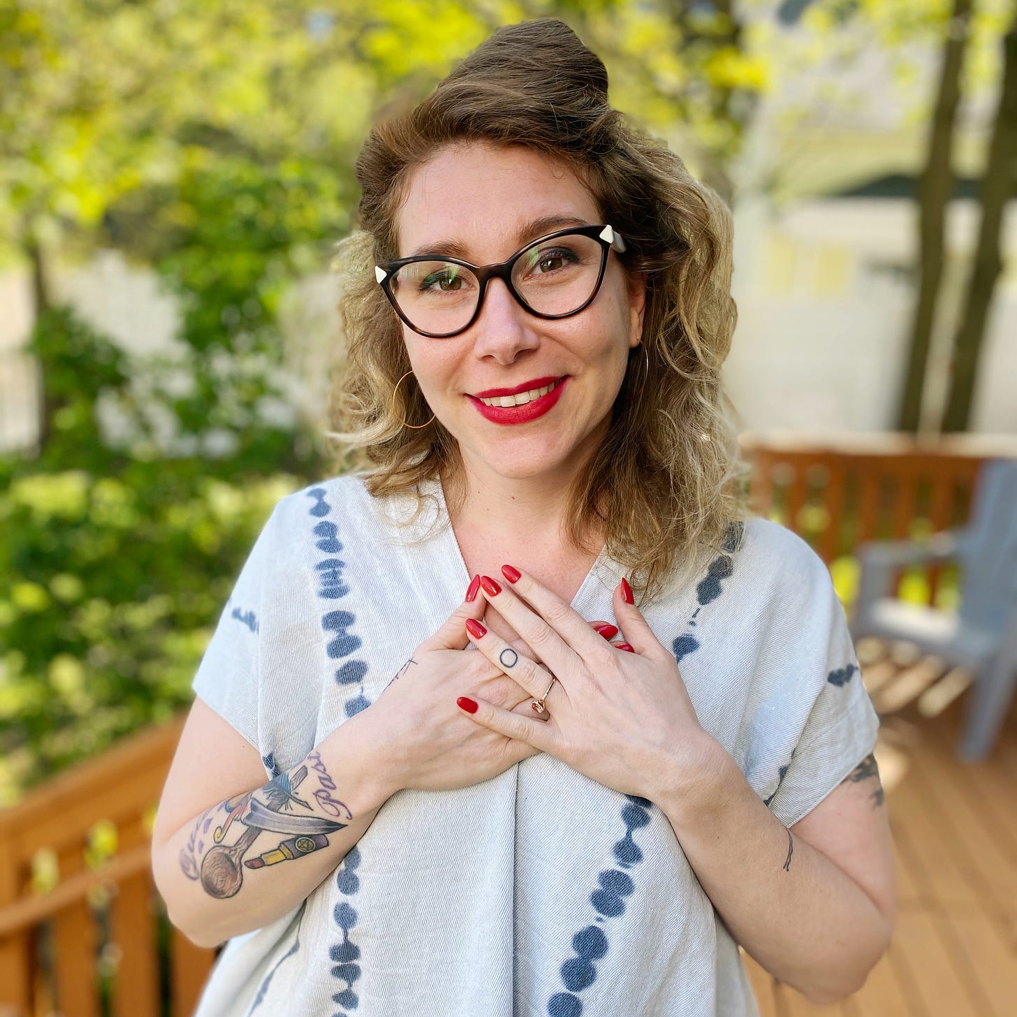 Dr. Kate is smiling at the camera. She is a white woman with red and blonde hair wearing a light blue shirt with dark blue died spots. She is wearing glasses, red lipstick, and red nails.