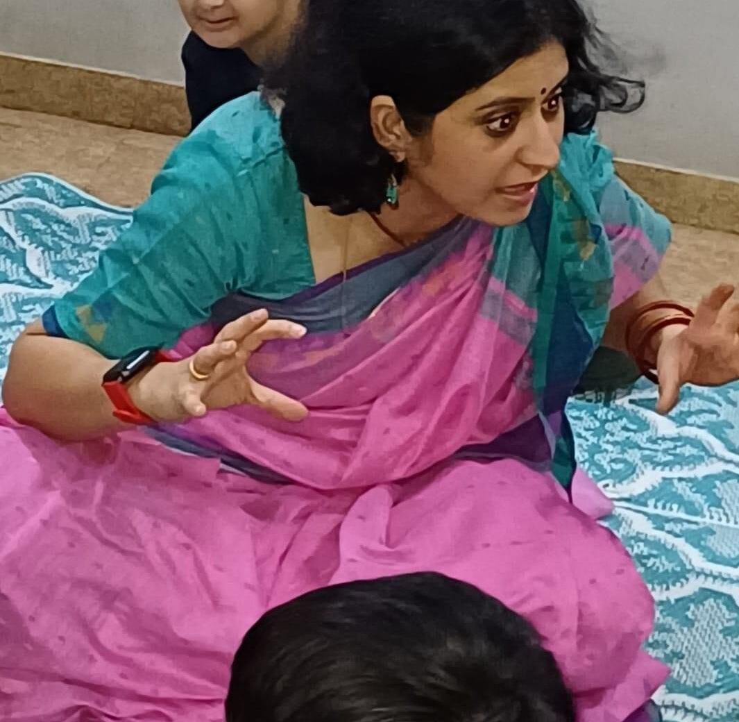 Dressed in a teal and magenta saree, Shivani is seated with folded legs on a blue and white patterned mat. She is an Indian woman with short black hair and her bindi, bangles, and watch are red. She is looking to her left, narrating a story to a group of kids, who are not in the frame. Shivani’s hands, lifted with fingers gently folded, mimic a lion ready to spring upon its prey. 