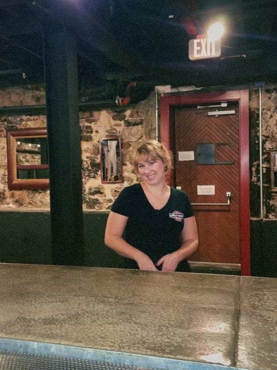 Hayley, a white blonde woman, stands behind a bar wearing a black t-shirt. Behind her is a speakeasy door and an old stone wall.