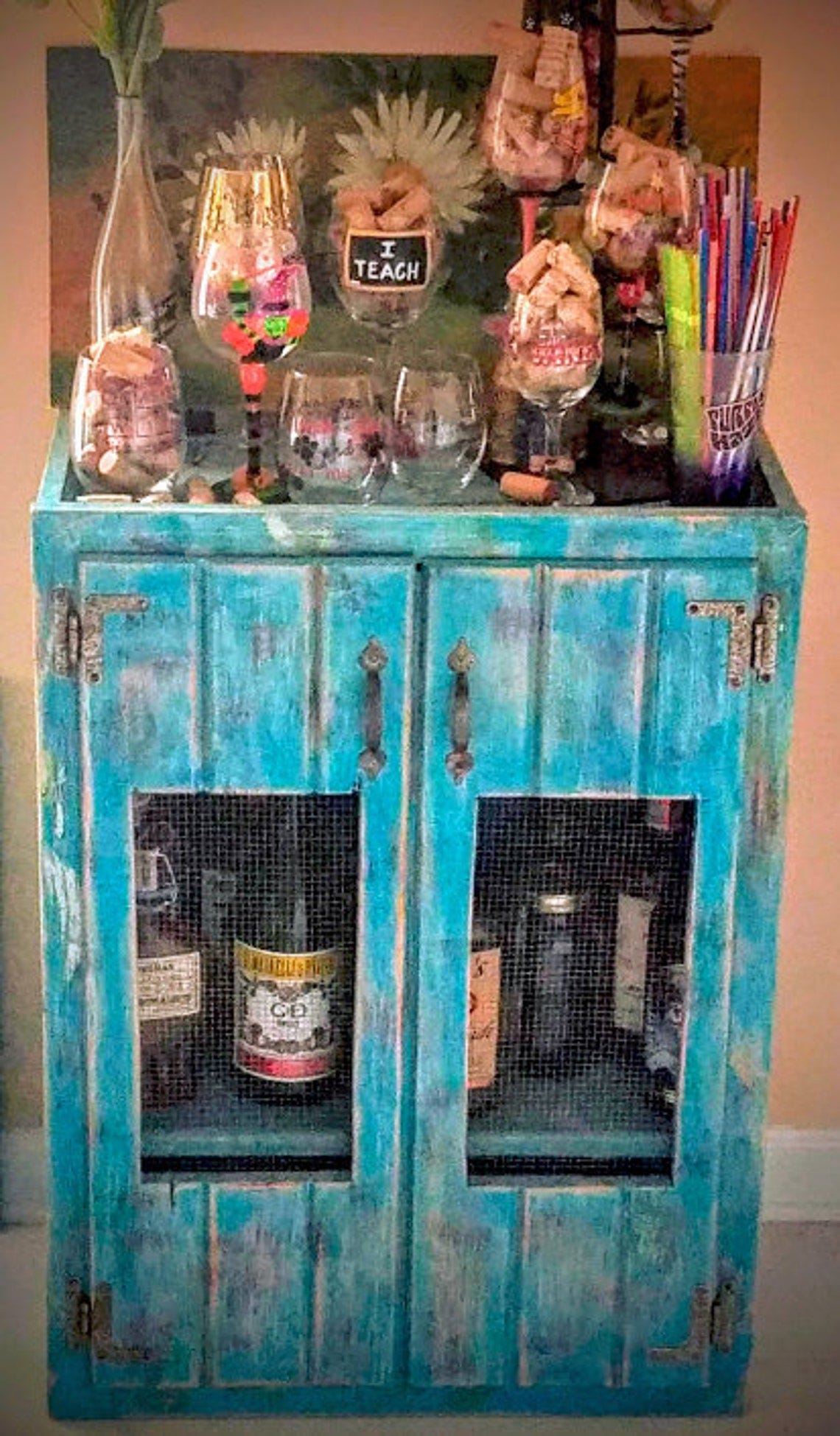 May include: A turquoise blue cabinet with two doors and glass panels. The cabinet has a distressed finish and is decorated with metal hinges and handles. The cabinet is filled with various bottles and glasses. A sign on the top of the cabinet reads 'I Teach'.