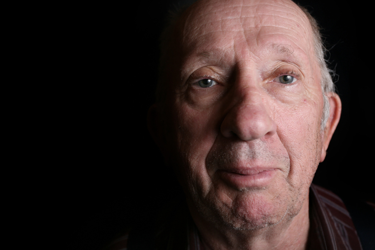 Elderly man without glasses looking at viewer. Light highlights right side of face. Black background. 