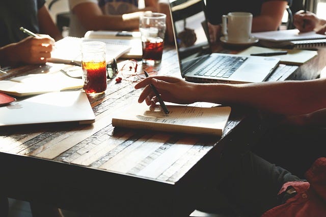 Photo of table with glimpses of people around it with pens poised over notebooks, surrounded by drinks and laptops in an informal meeting