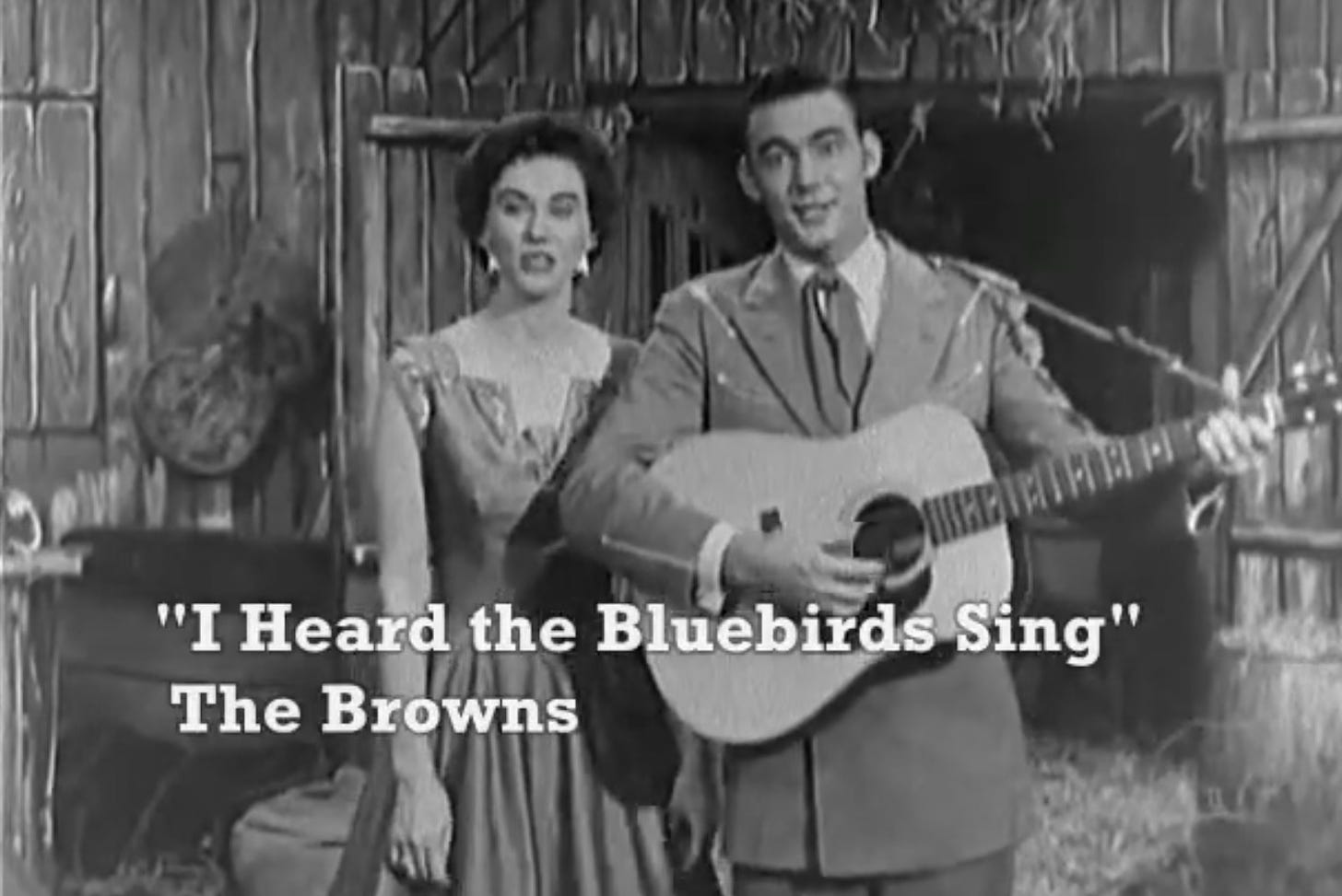 The Browns singing the song I Heard the Bluebirds Sing in front of an old homestead building. Maxine Brown is on Jim's left as he strums a guitar