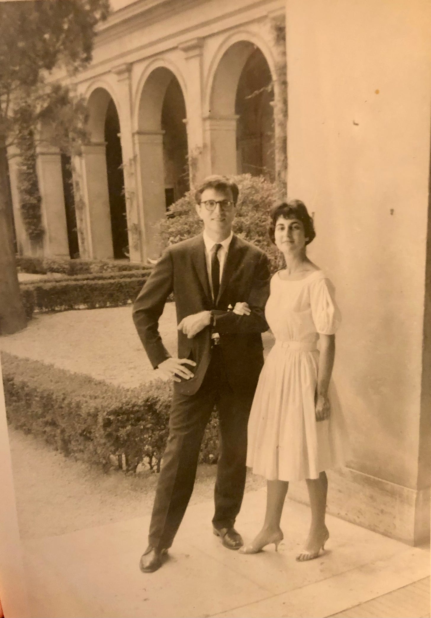 A couple dressed for a party pose for a photo, her arm in his, in a courtyard with archways behind them and hedges.