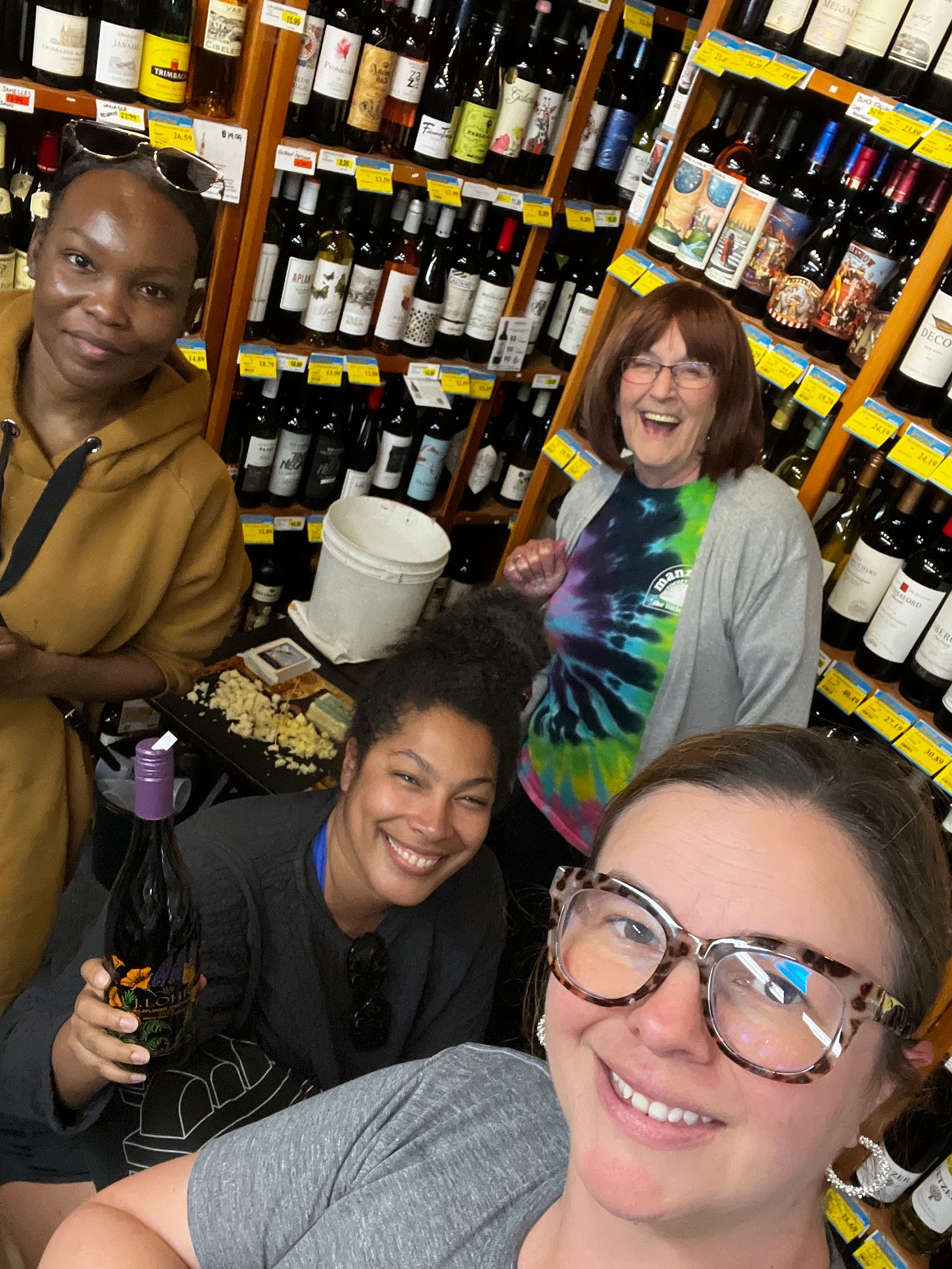 Nafissa, Jan, Jackie, and Amber take a group selfie together in a grocery store. Behind them, shelves of wine bottles can be seen as well as a small table for wine tasting. Jackie holds a wine bottle up. Everyone smiles.