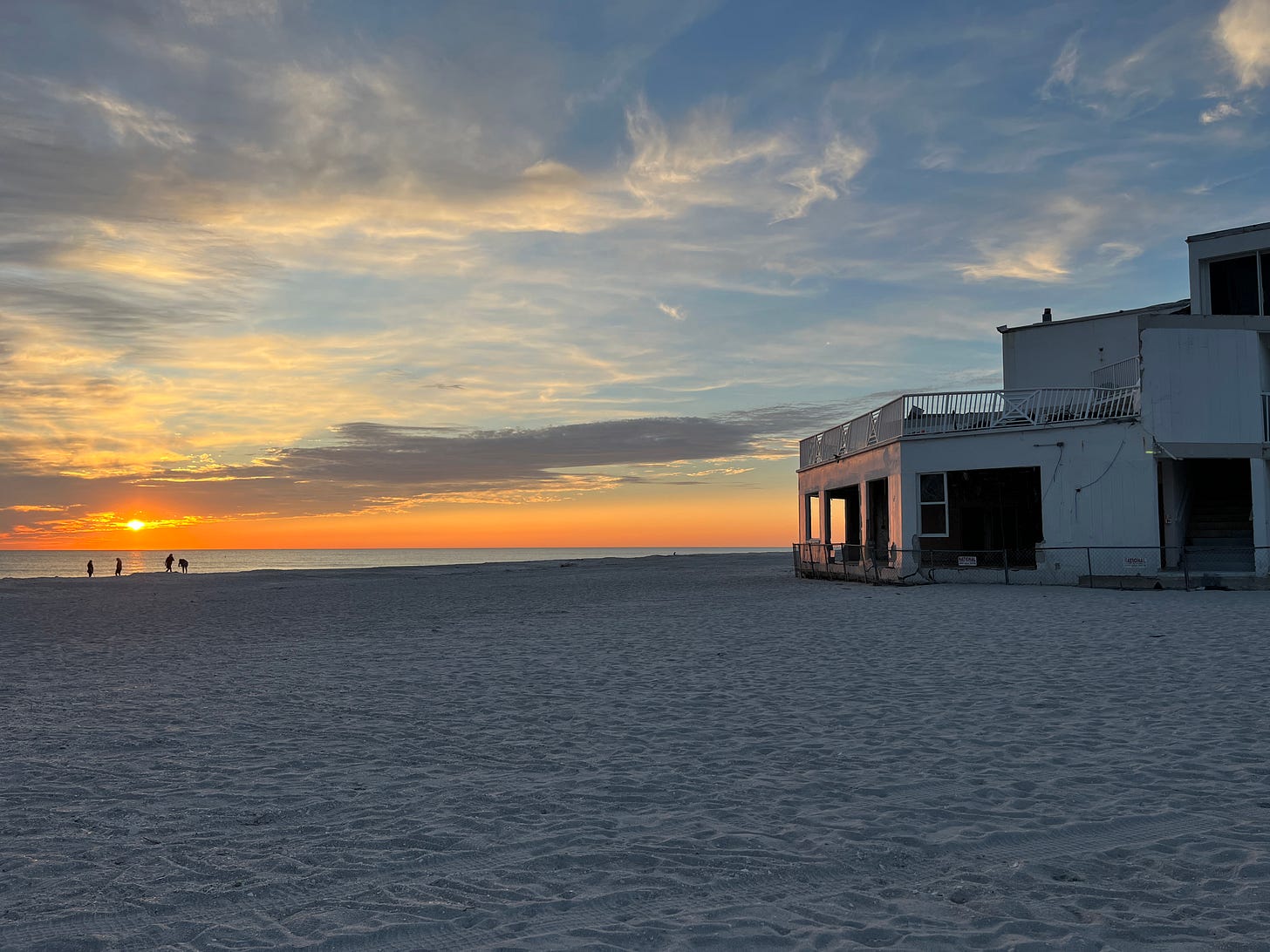 The edge of a building that is clearly empty, with no windows and debris hanging off of it, with a spectacular sunset off to the side and some figures at the water line