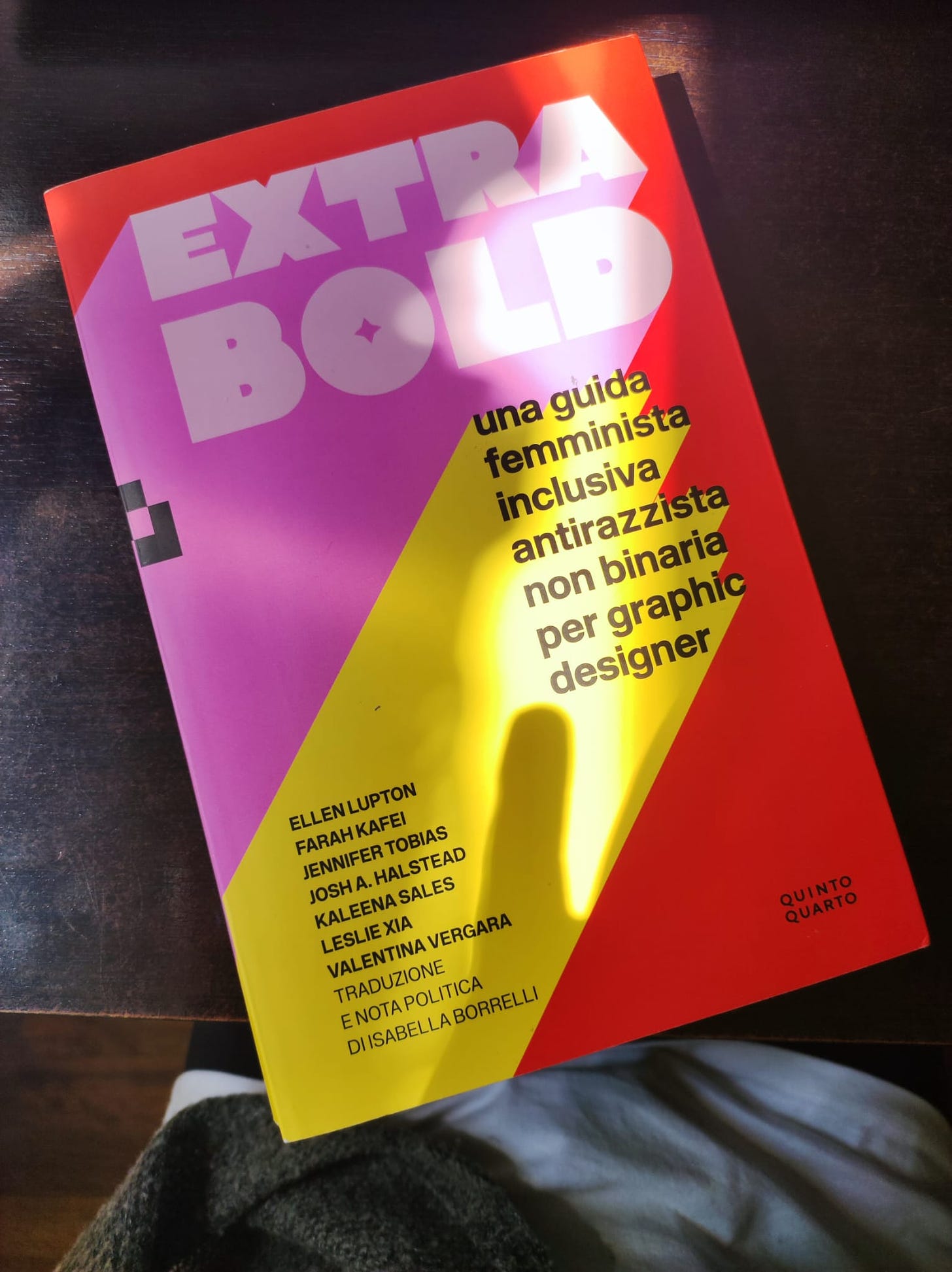 Libro appoggiato su una scrivania scura. La copertina del libro, Extra Bold, ha composizioni geometriche colorate di rosso, rosa e giallo.