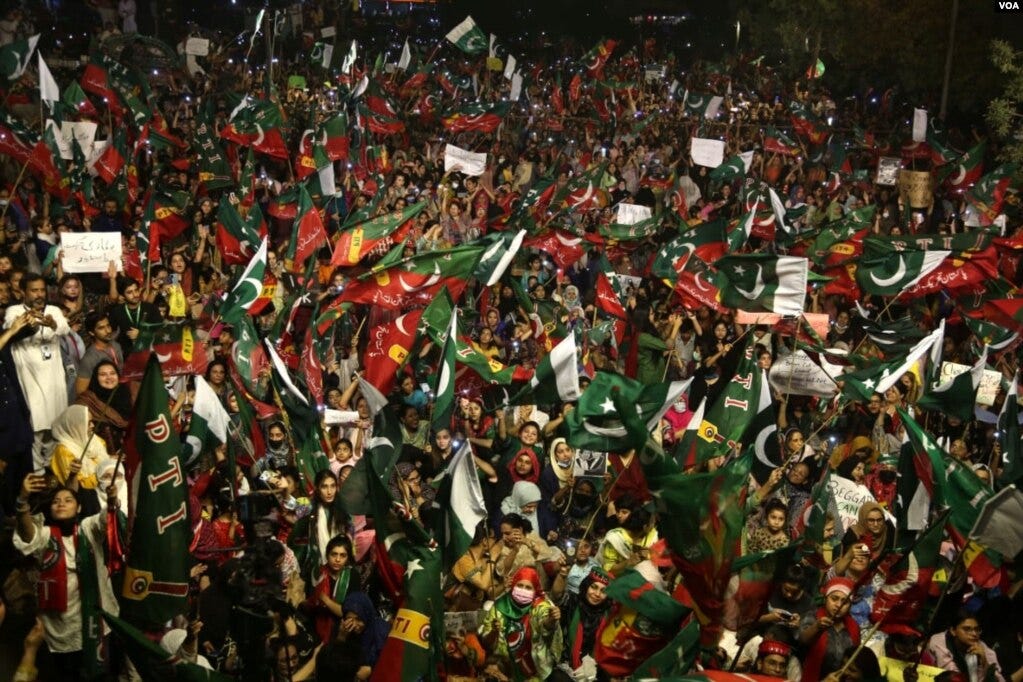 A massive crowd of people waving red and green flags with a white crescent, showing support for the PTI.