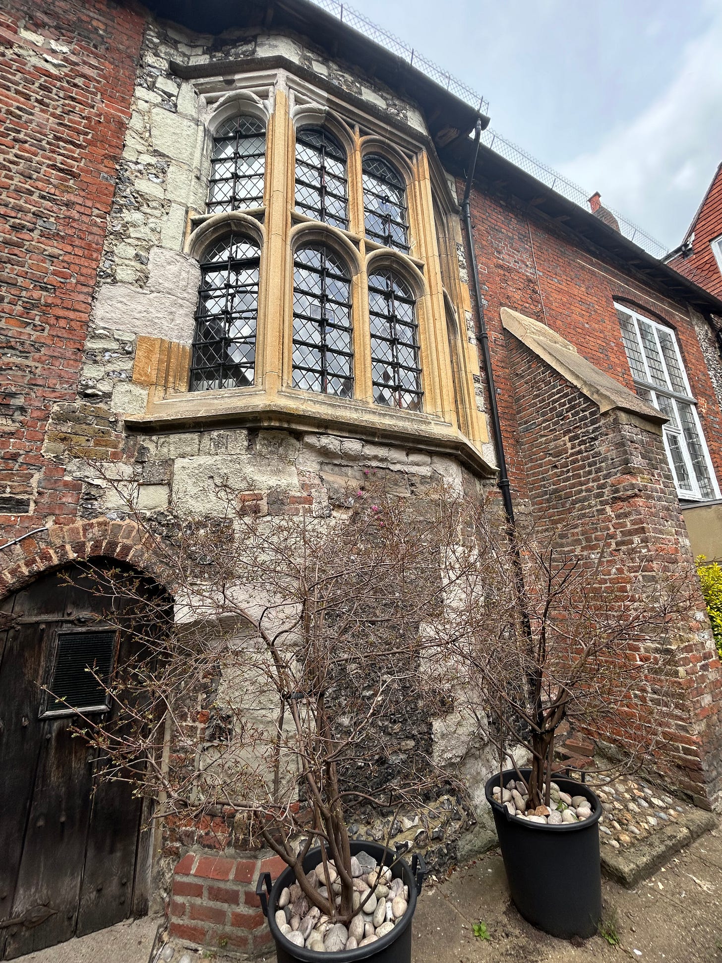 An old building with a large oriel window. The brick is uneven and clearly centuries old - some is gray stone, other areas are red brick with Tudor style construction.