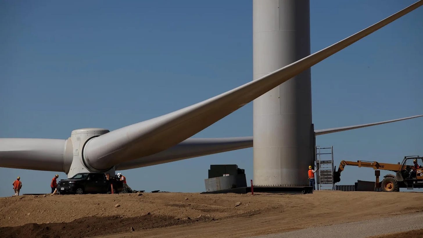 A photo of a wind turbine rotor and blades sitting on the ground next to an erected wind turbine tower, with the diminutive figures of workers dressed in red indicating the huge scale of the turbine.