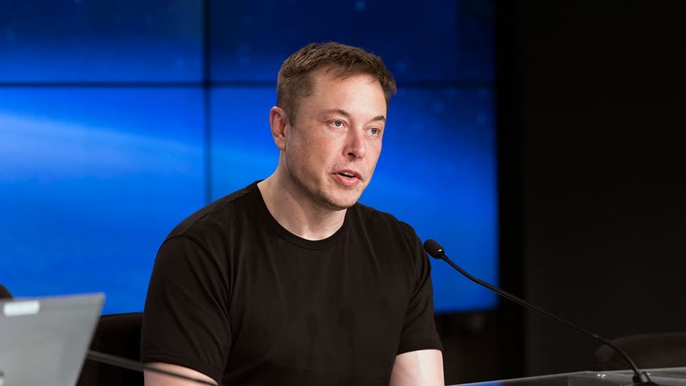 Elon Musk wearing a black t-shirt and talking into a microphone in front of some blue screens
