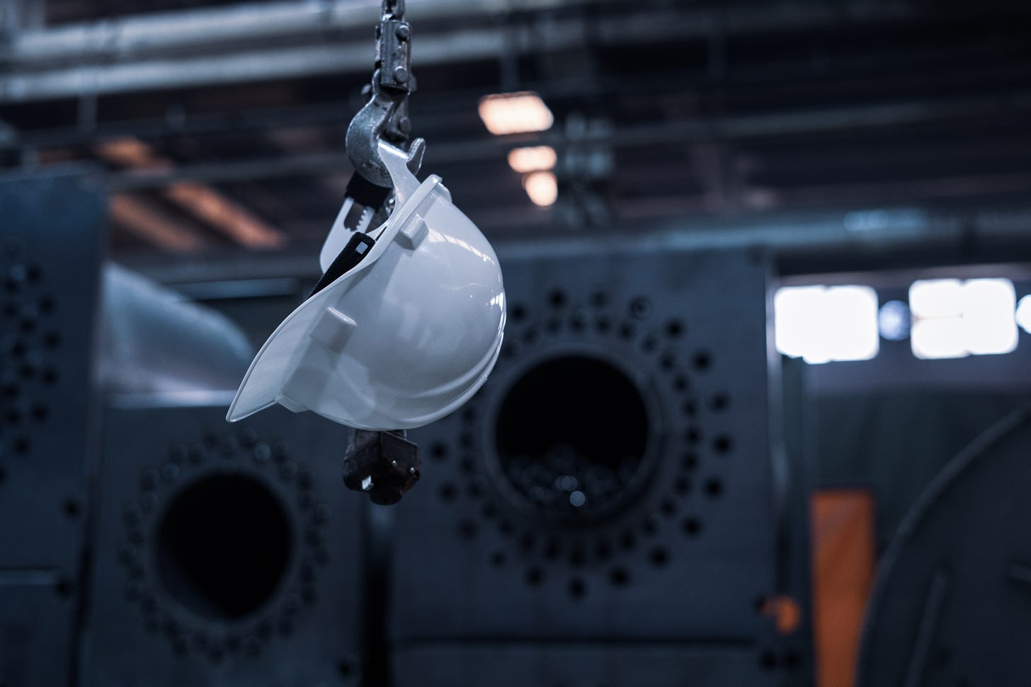 A white safety helmet hangs from a metal hooks, with industrial equipment out-of-focus in the background