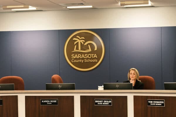 Bridget Ziegler sits by herself at a long desk used for meetings of the Sarasota County School Board. Nameplates for each board member are arranged along the front of the desk, and the logo of Sarasota County Schools hangs on the wall behind her.